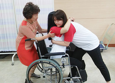 휠체어 사용자를 돕는 법을 연습하고 있는 학생과 지도하는 교수님. 한 학생이 환자의 연기를 하고 있다.