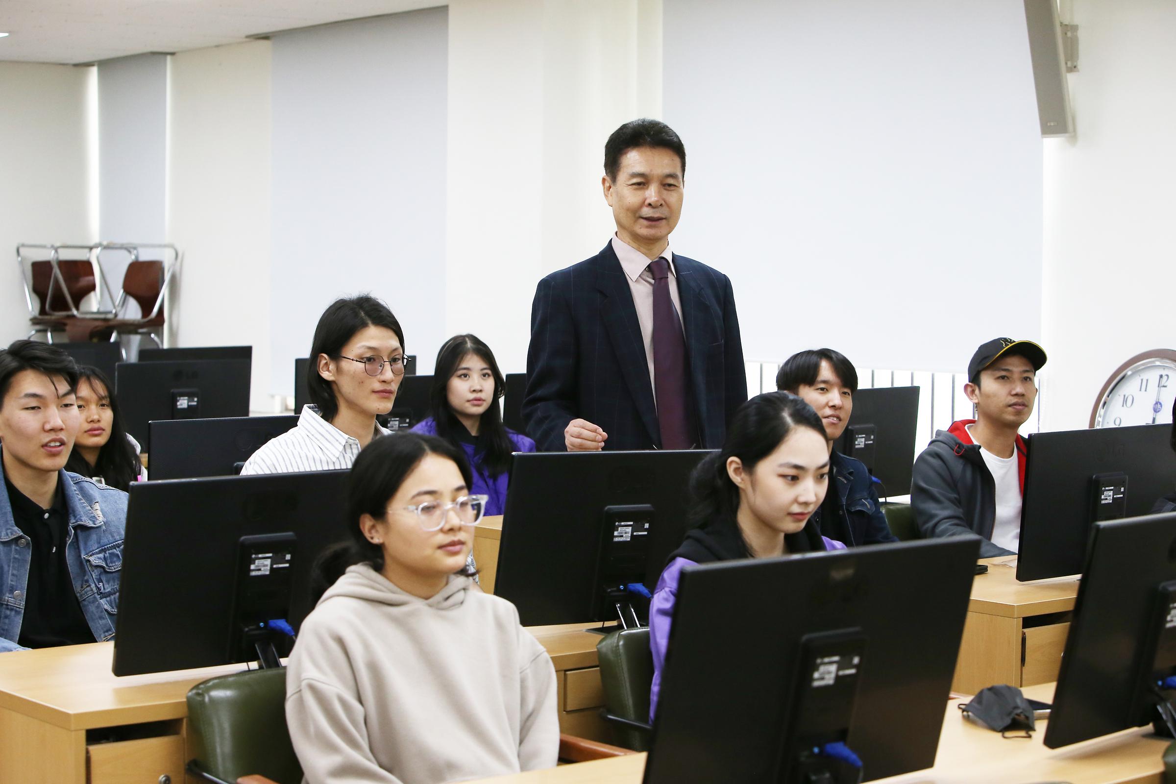 컴퓨터 실에서 수업을 듣는 학생들의 사진. 두 줄의 책상 사이에 교수님이 서있다.