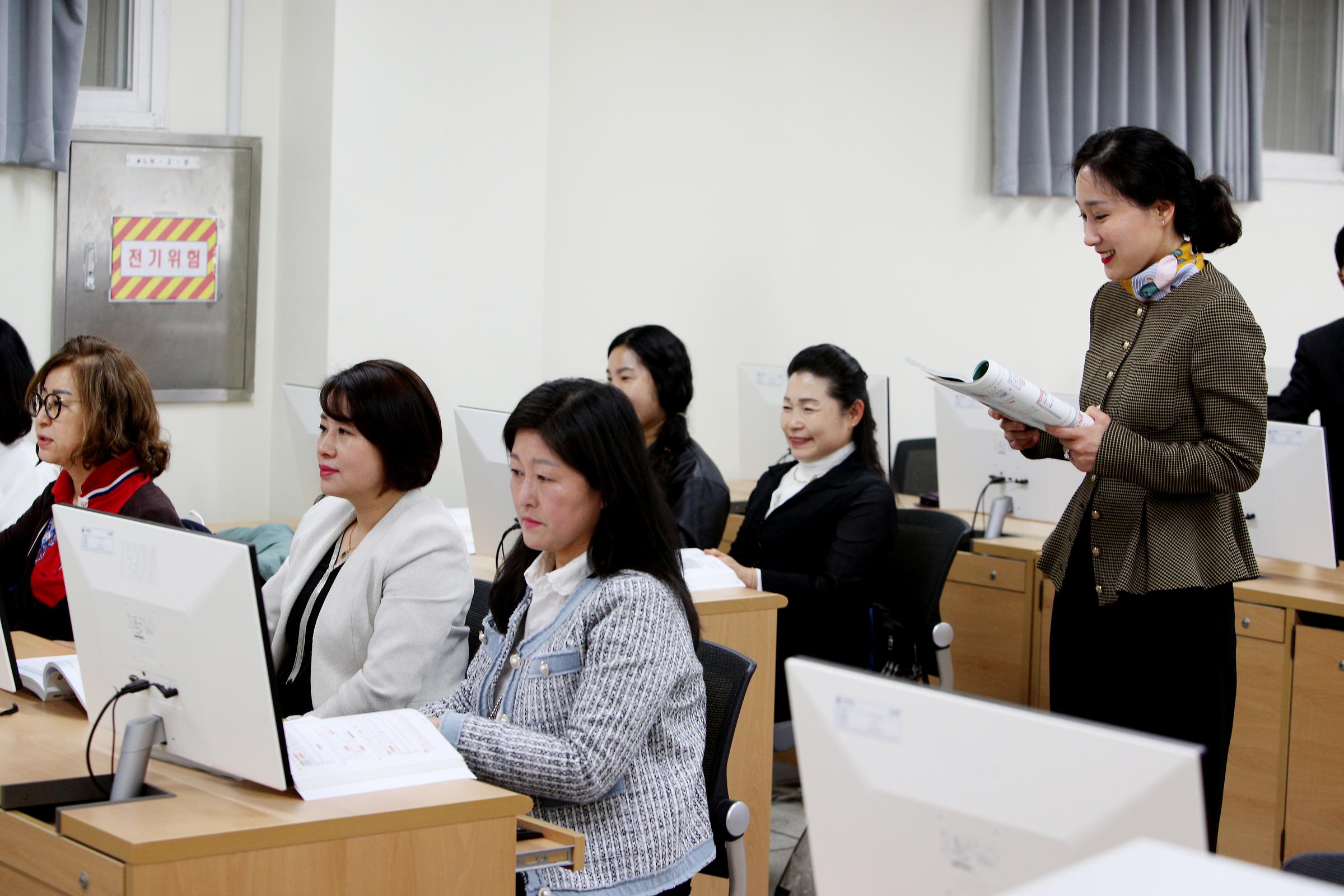 컴퓨터실에서 수업을 듣는 학생들의 사진. 중년의 여학생들이 컴퓨터 앞에 앉아있고 교수님이 서서 수업중이다.